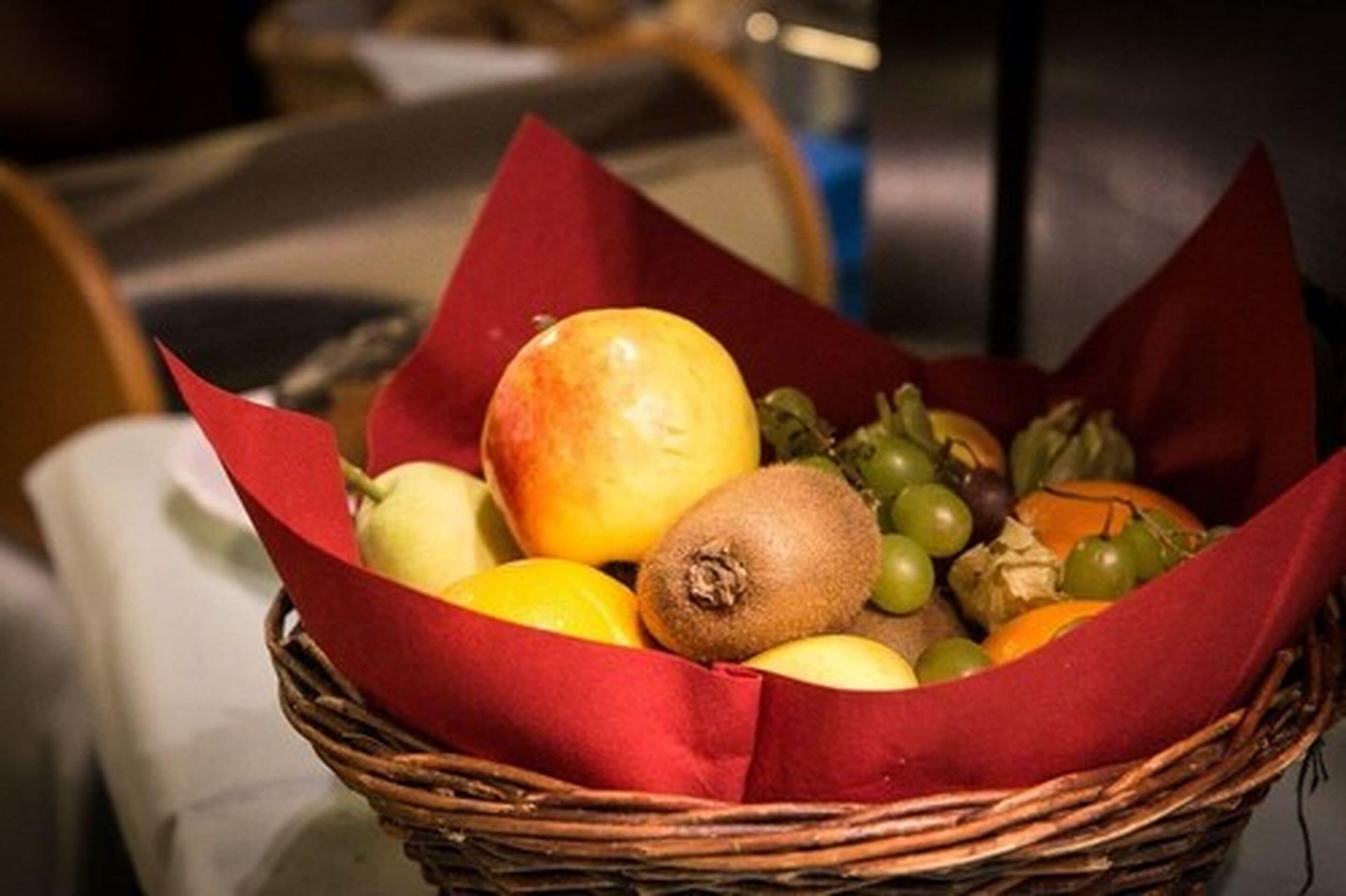 Una cesta de mimbre llena de frutas y verduras, como manzanas, kiwis y uvas, envueltas en papel rojo.
