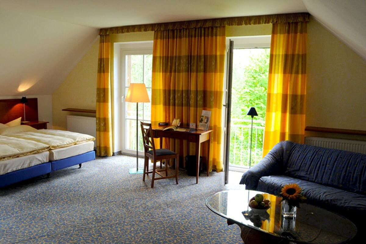 Habitación de hotel con cama, escritorio con silla, sofá y balcón con cortinas.
