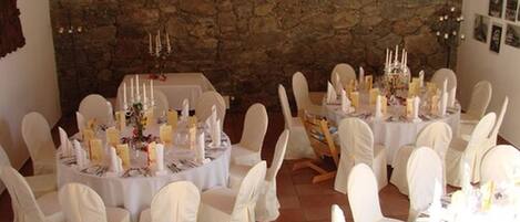 Un salón de banquetes con mesas redondas dispuestas para un evento formal, sillas blancas y un fondo de pared de piedra.
