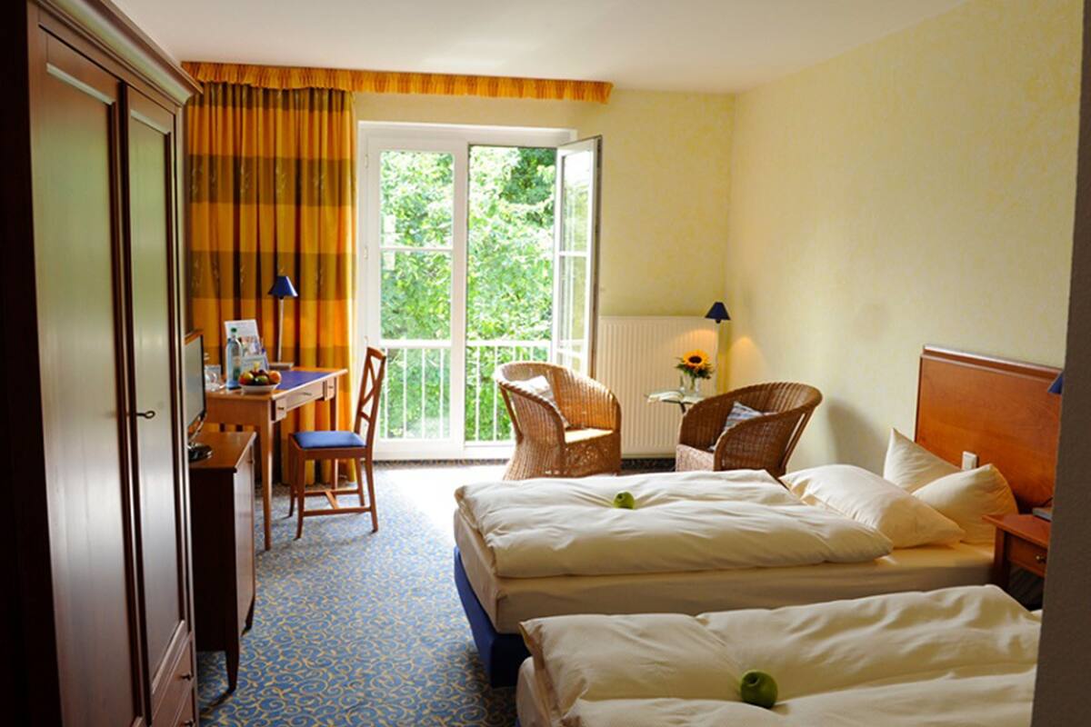 Habitación de hotel con dos camas, un escritorio con silla, una ventana con cortinas y vistas a zonas verdes.