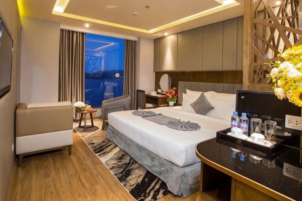 A hotel room with a bed, a desk, a chair, a sofa, a TV, and a view of the outside.