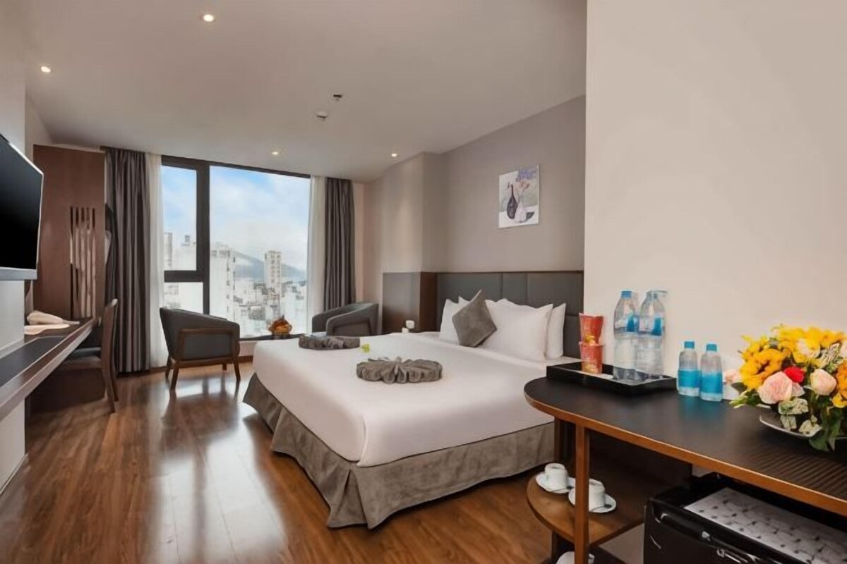 A modern hotel room with a large bed, a desk, a chair, and a view of the city.