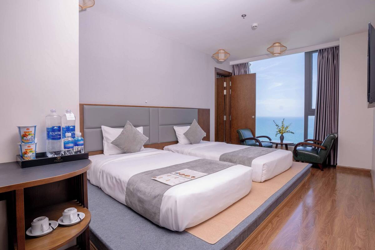 Ein Hotelzimmer mit zwei Betten, einem kleinen Tisch mit Frühstücksangeboten und Meerblick, sowie einem großen Fenster mit Vorhängen.