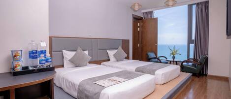 Quarto de hotel com duas camas, uma pequena mesa com itens de café da manhã, vista para o mar e uma janela grande com cortinas.