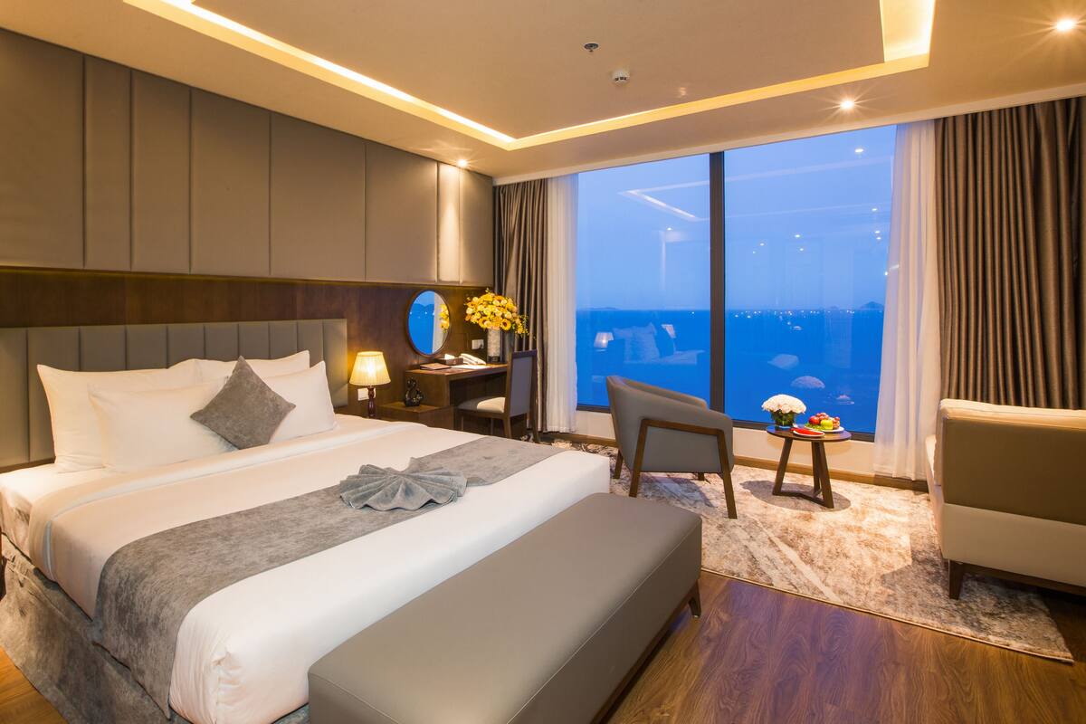 Ein Hotelzimmer mit einem großen Bett, einem Schreibtisch, einem Sessel und Blick aufs Meer.