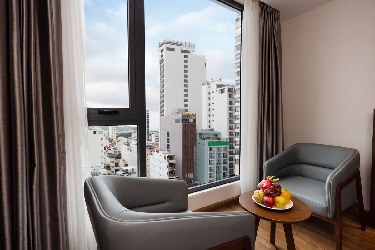 Ein modernes Hotelzimmer mit einem großen Fenster, zwei grauen Sesseln und einem kleinen Tisch mit Obstschale.