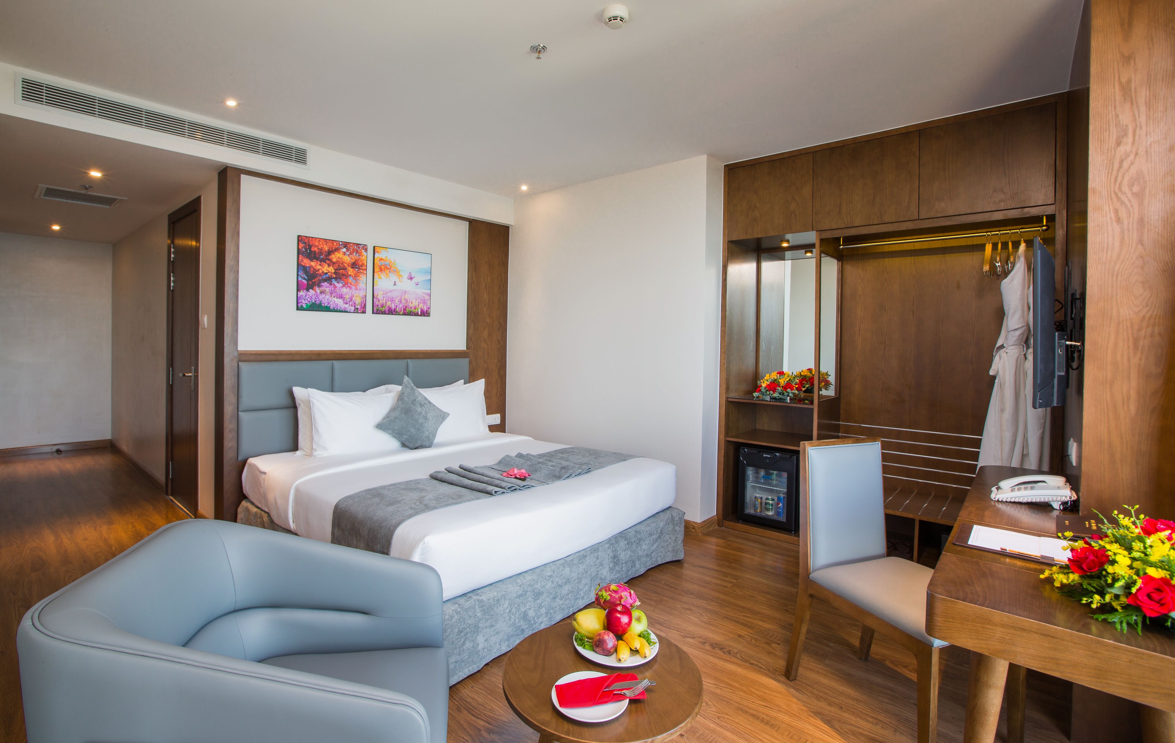 A modern hotel room with a bed, a seating area with a table, a chair, and a small dining table with a fruit bowl.