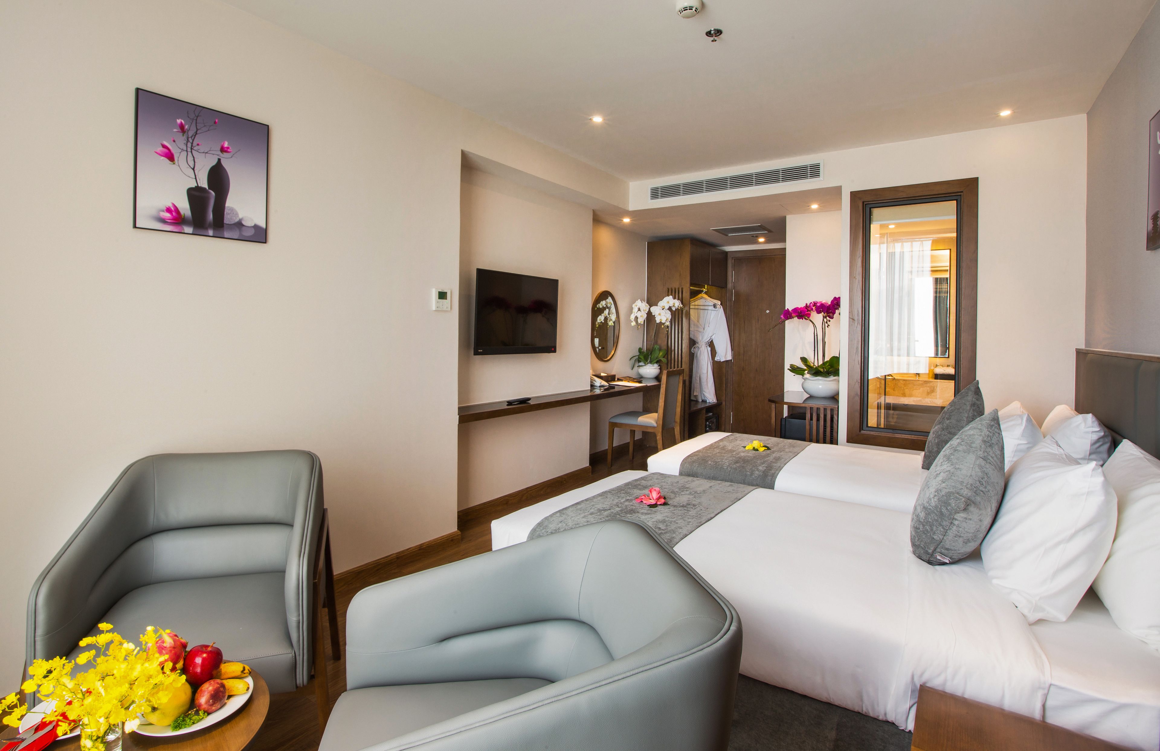 A hotel room with a bed, two armchairs, a TV, a mirror, a desk, and a bathroom visible through an open door.