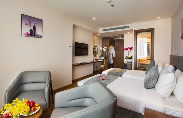 A hotel room with a bed, two armchairs, a TV, a mirror, a desk, and a bathroom visible through an open door.