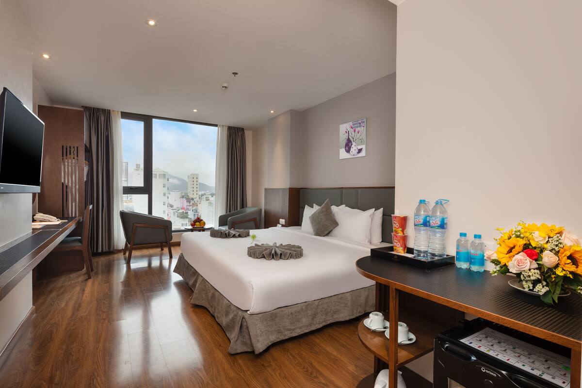 Ein Hotelzimmer mit einem großen Bett, einem Schreibtisch, einem Sessel, einem Fernseher und Blick auf die Stadt.