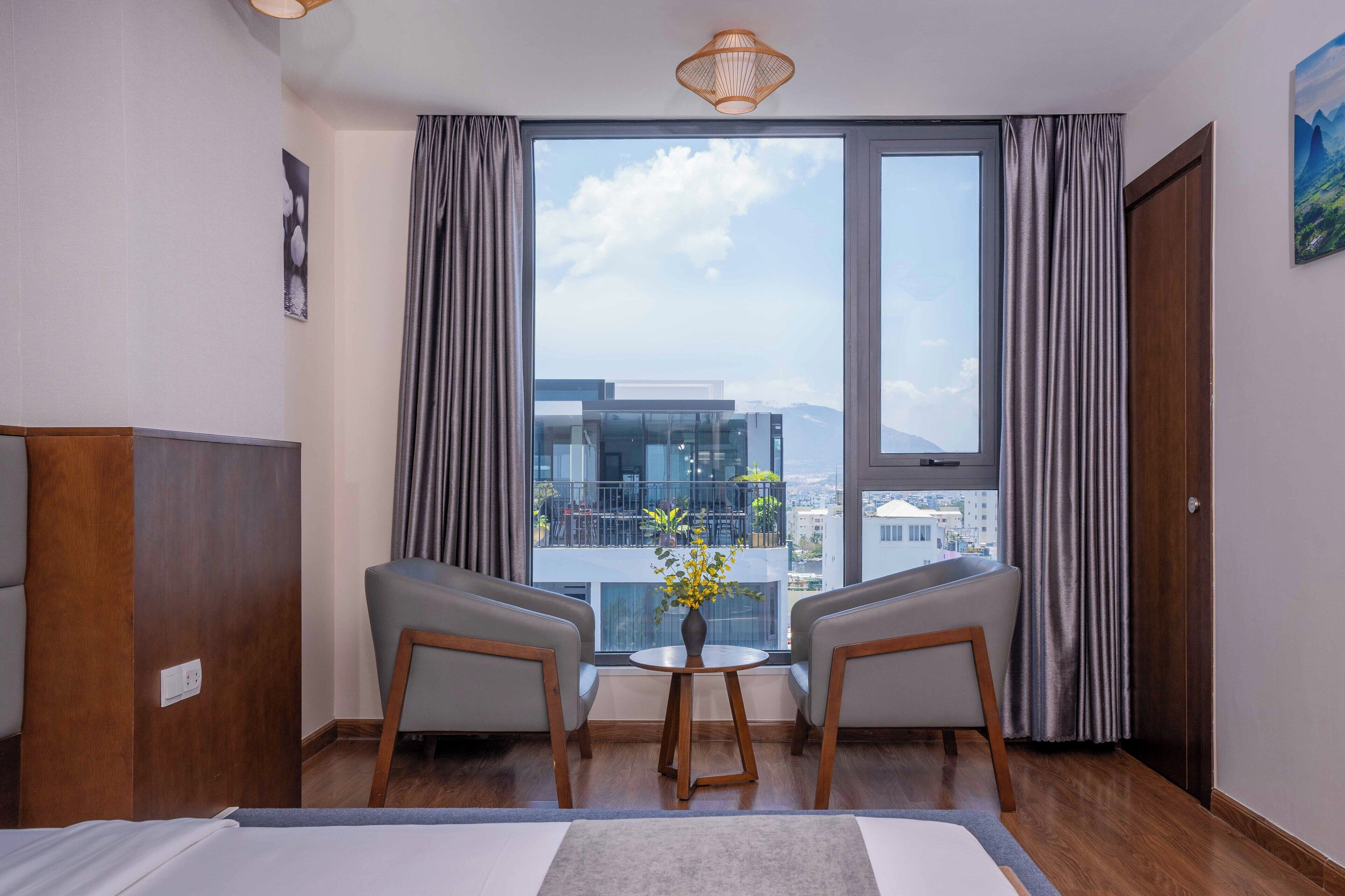 A hotel room with a large window, two armchairs, a small table with a vase, and a bed.