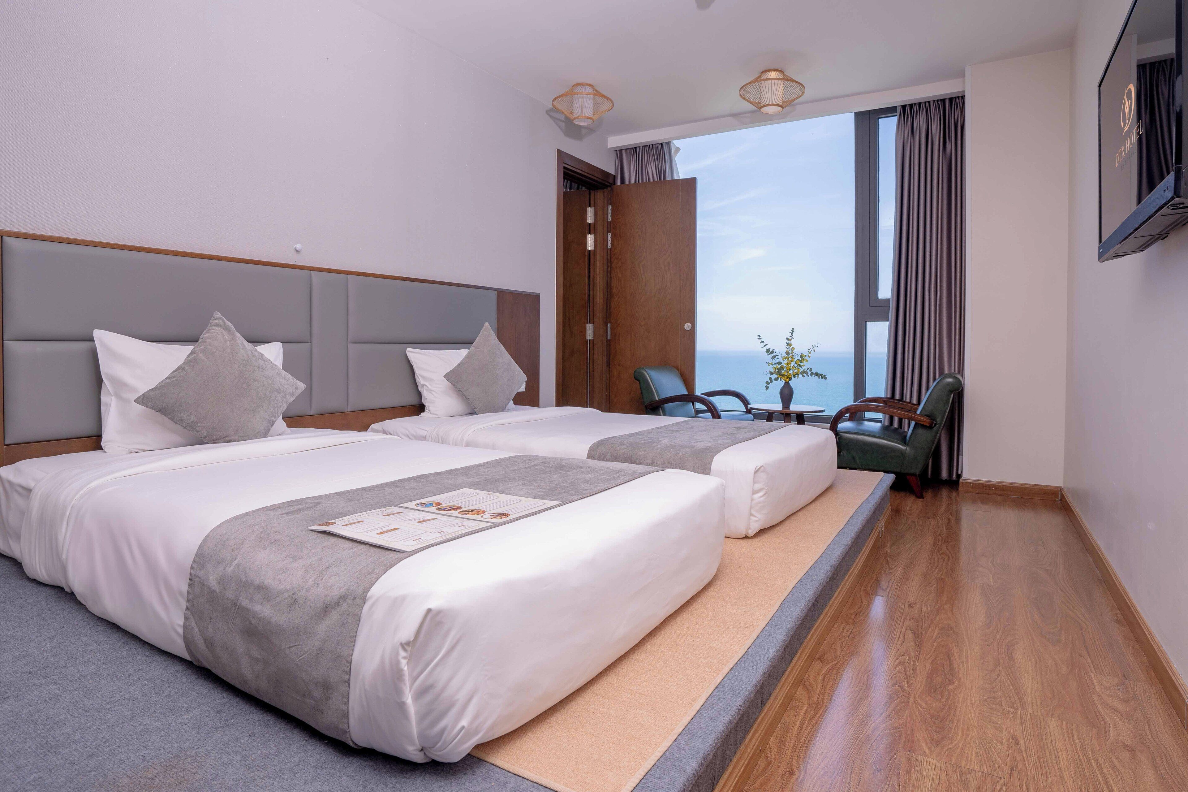 A hotel room with two beds, a wooden headboard, a large window with curtains, a TV, and a seating area with a chair and a table.