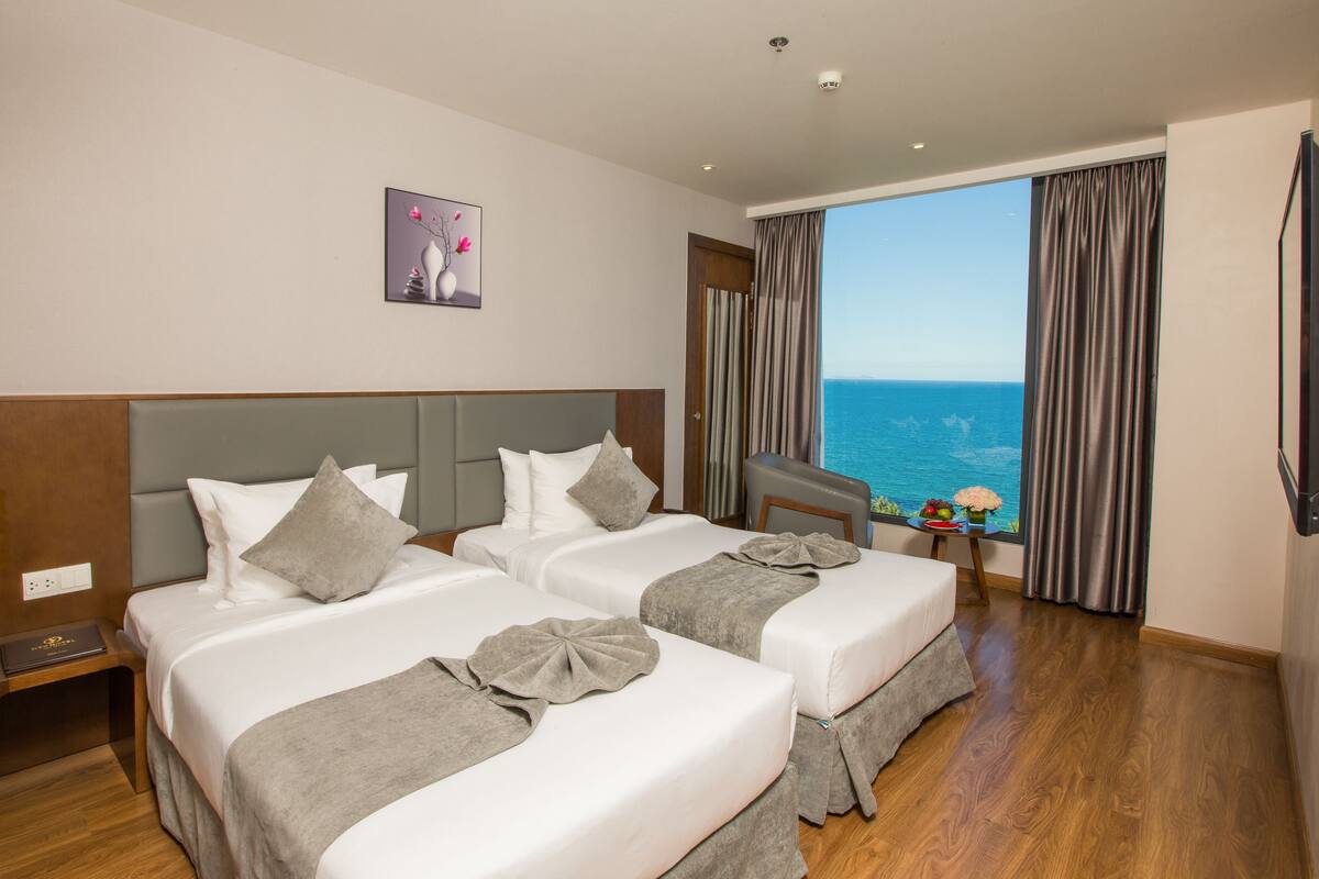 Ein Hotelzimmer mit zwei Betten, einem großen Fenster mit Meerblick, einem kleinen Tisch mit Blumen und einem Bild an der Wand.