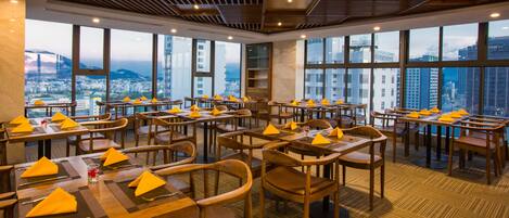 A modern restaurant with multiple tables and chairs, large windows offering city views, and a wooden ceiling.