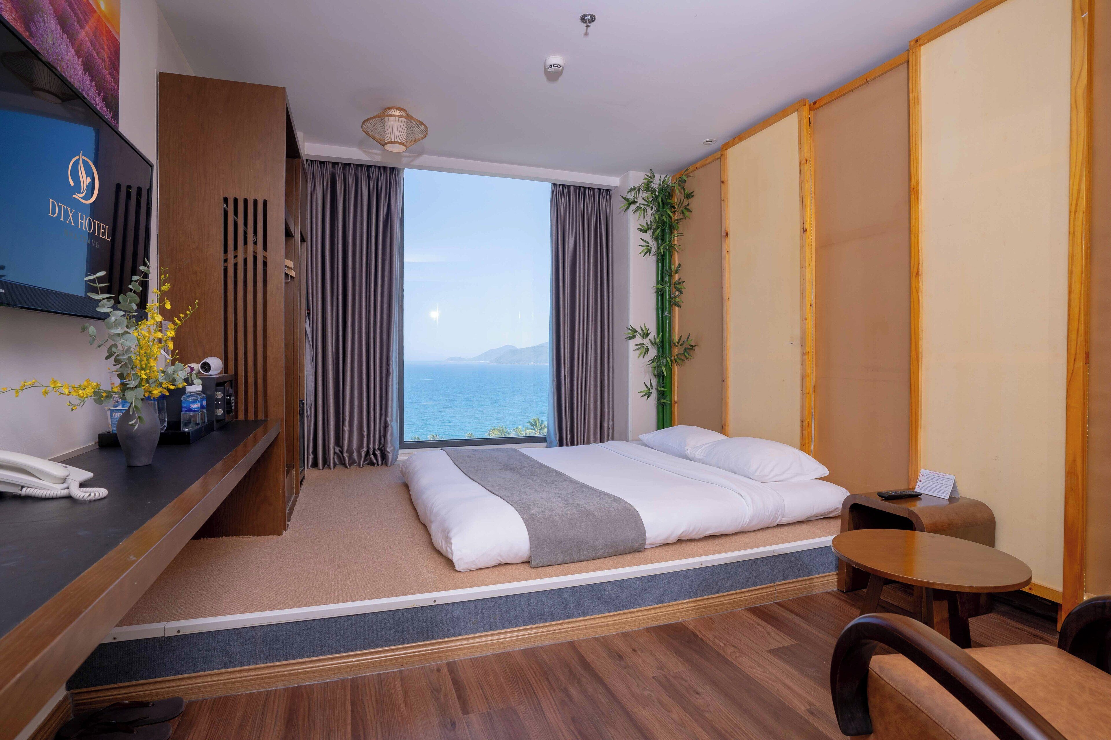 A hotel room with a bed, a desk, a TV, and a view of the sea.