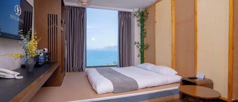 A hotel room with a bed, a desk, a TV, and a view of the sea.