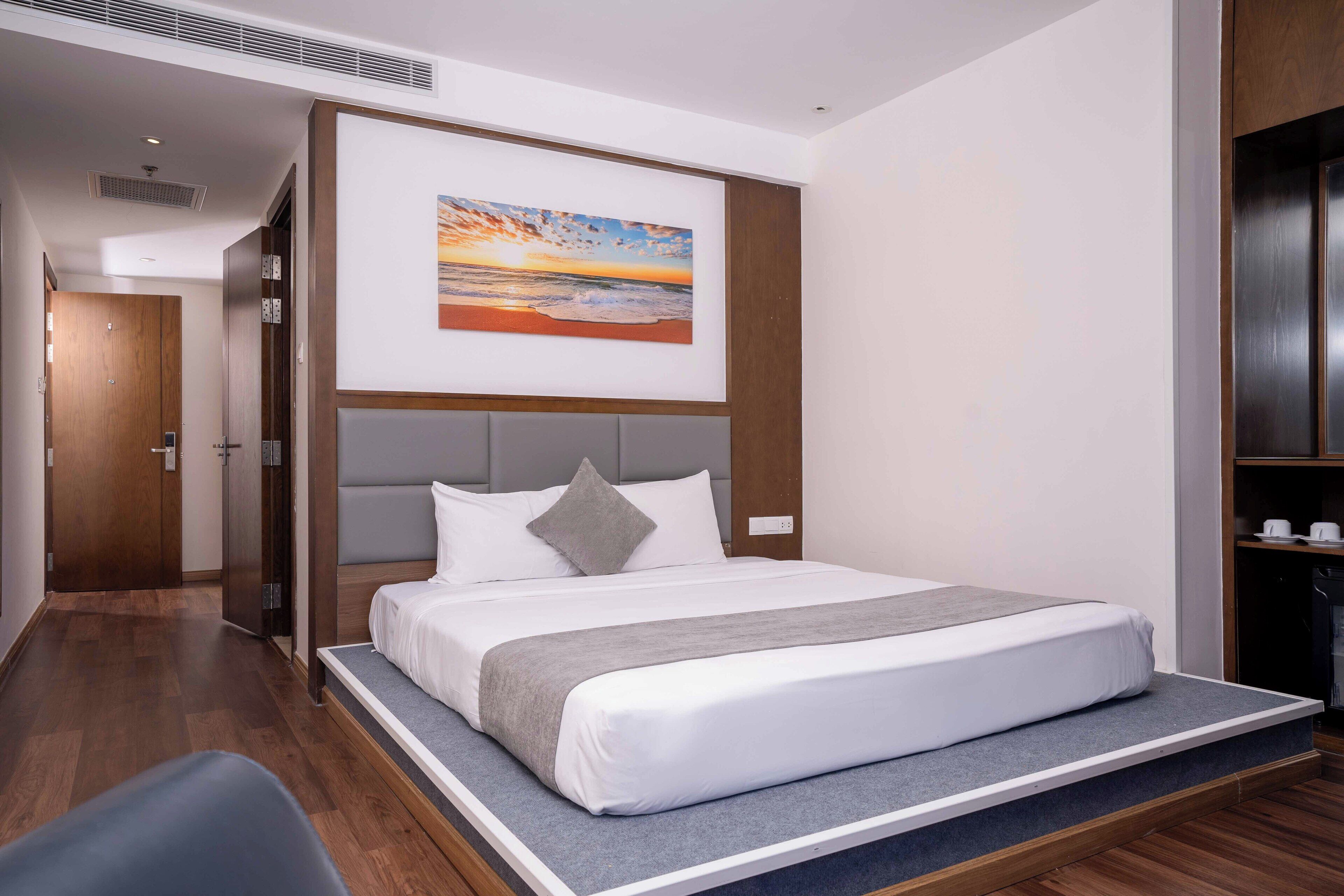 Quarto de hotel moderno com uma cama grande, cabeceira com prateleira embutida, pintura de um pôr do sol e piso de madeira.