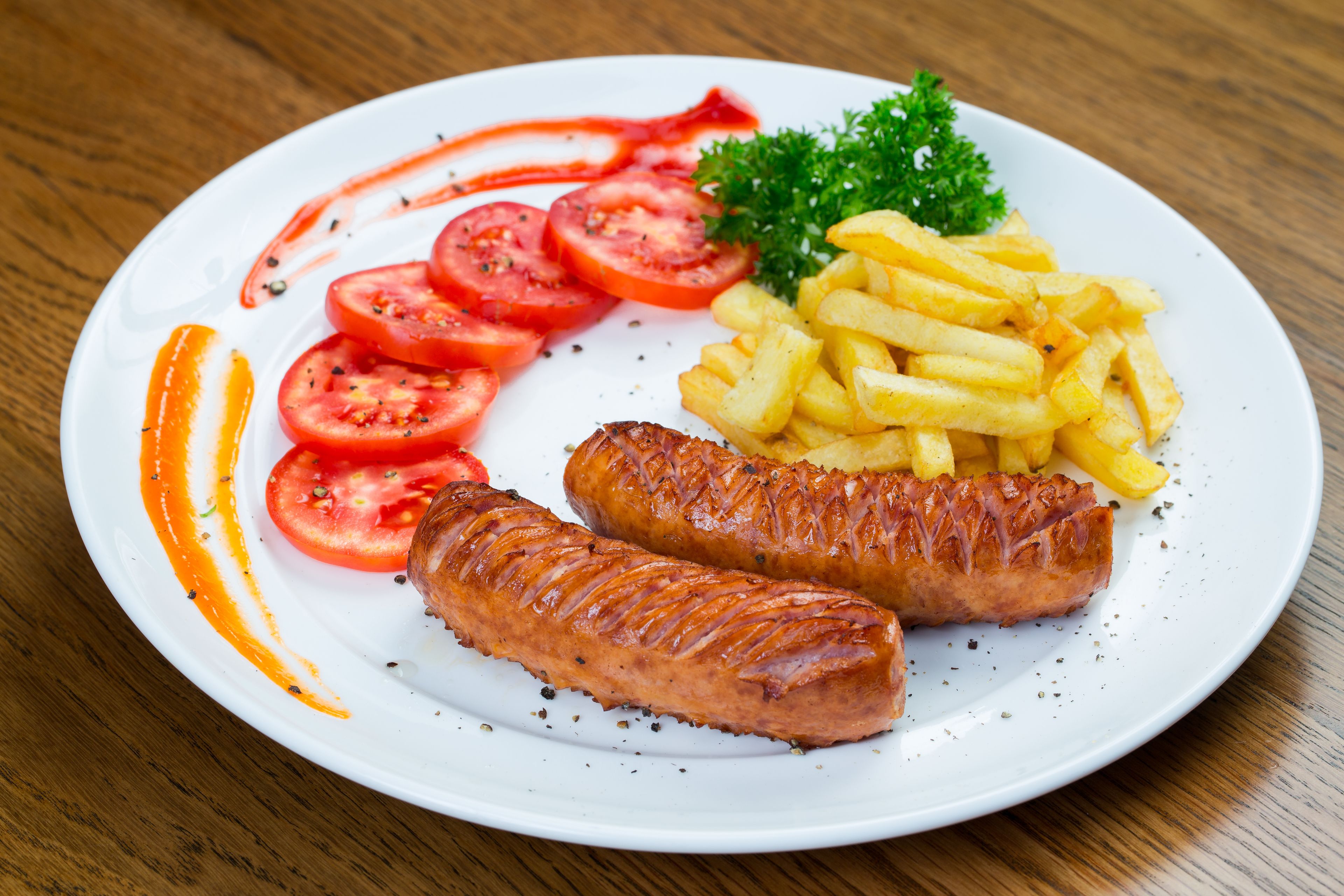 A plate with two grilled sausages, French fries, and sliced tomatoes on a white plate.