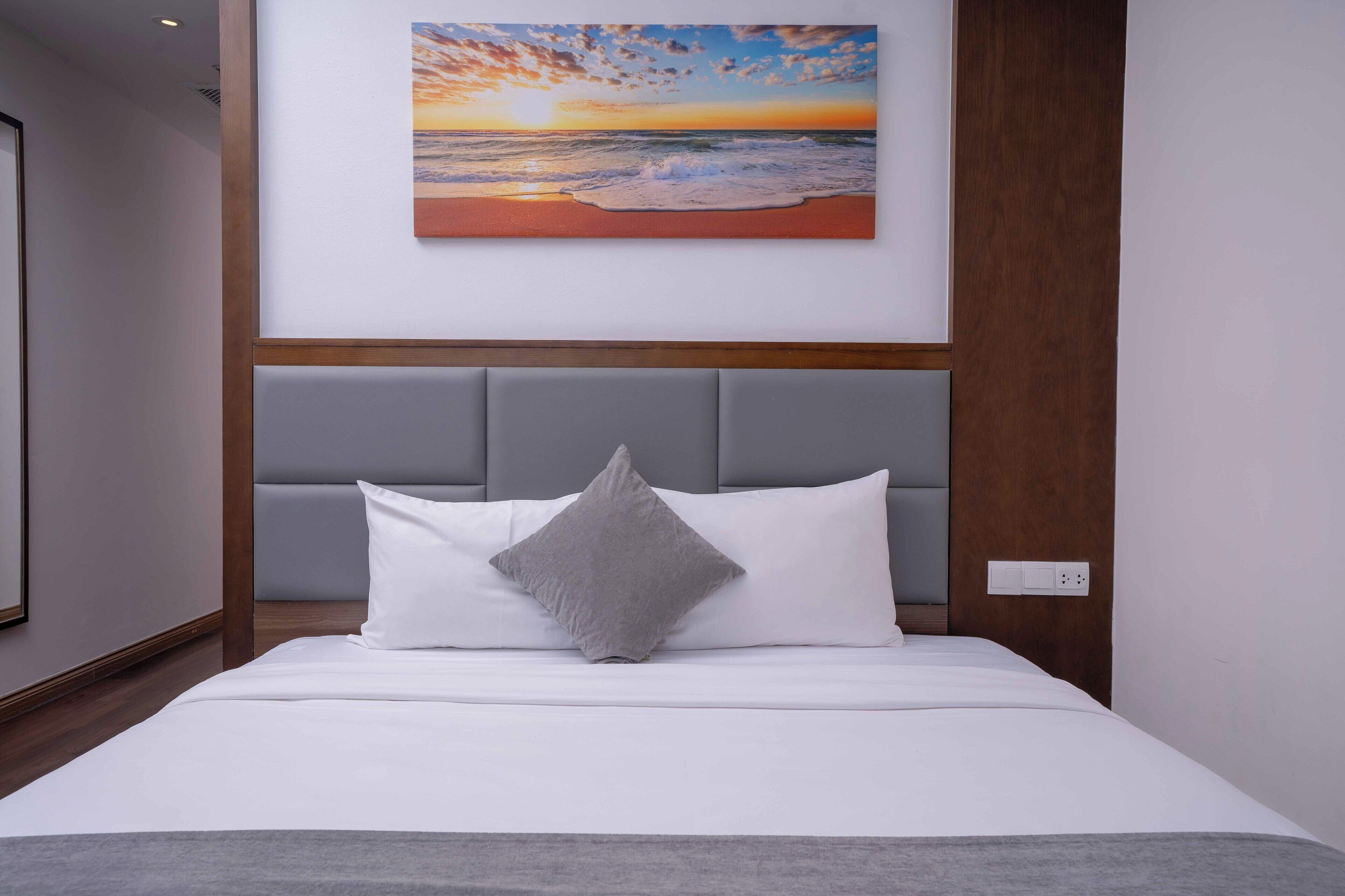 A neatly made bed with white bedding and a gray pillow, a gray headboard, and a painting of a sunset over the ocean on the wall above.
