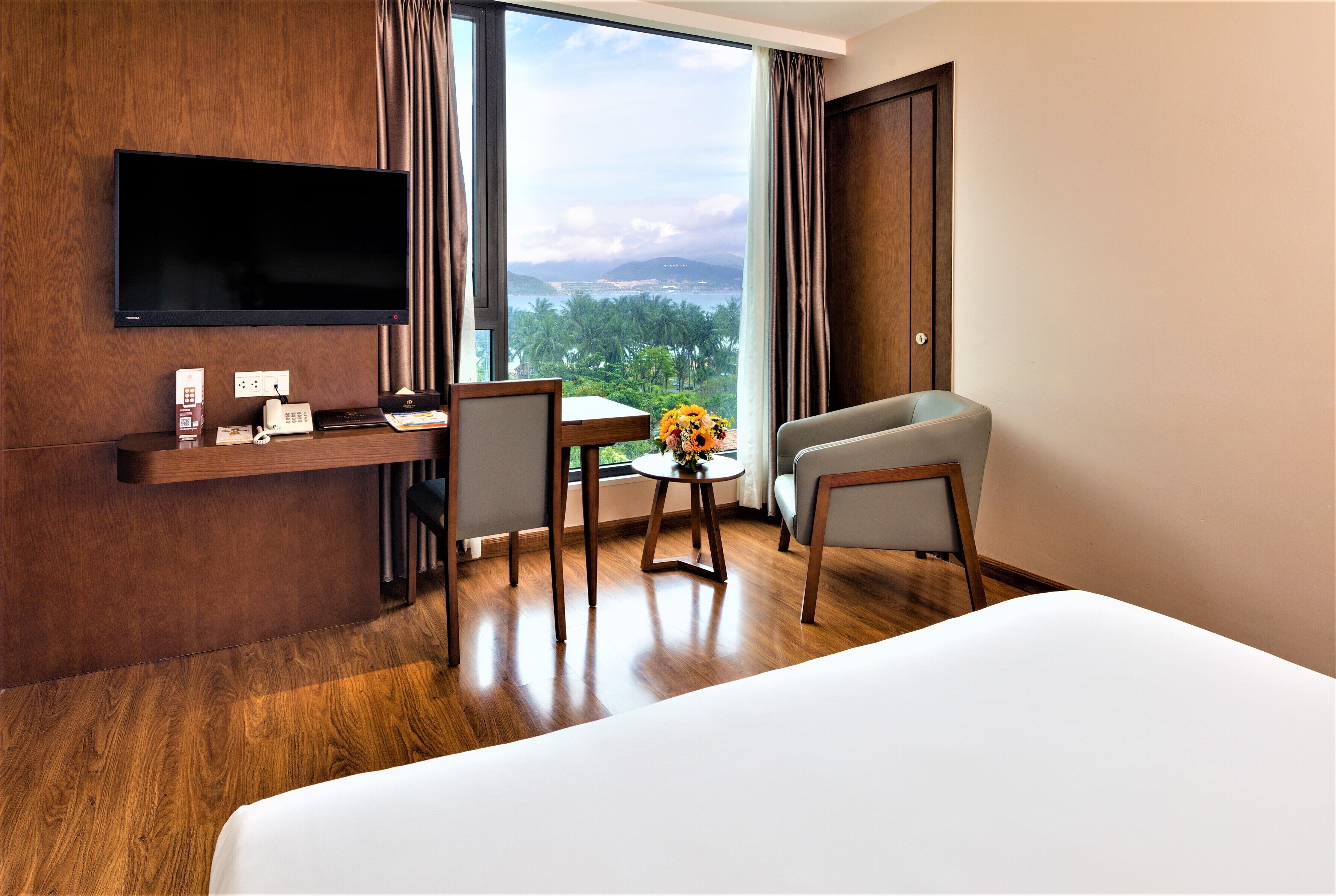 A hotel room with a bed, a desk with a flat-screen TV, a chair, a small table with a vase, and a view of the ocean and palm trees.