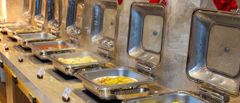 Buffet com diversos alimentos em recipientes de metal, alguns com detalhes vermelhos no topo.
