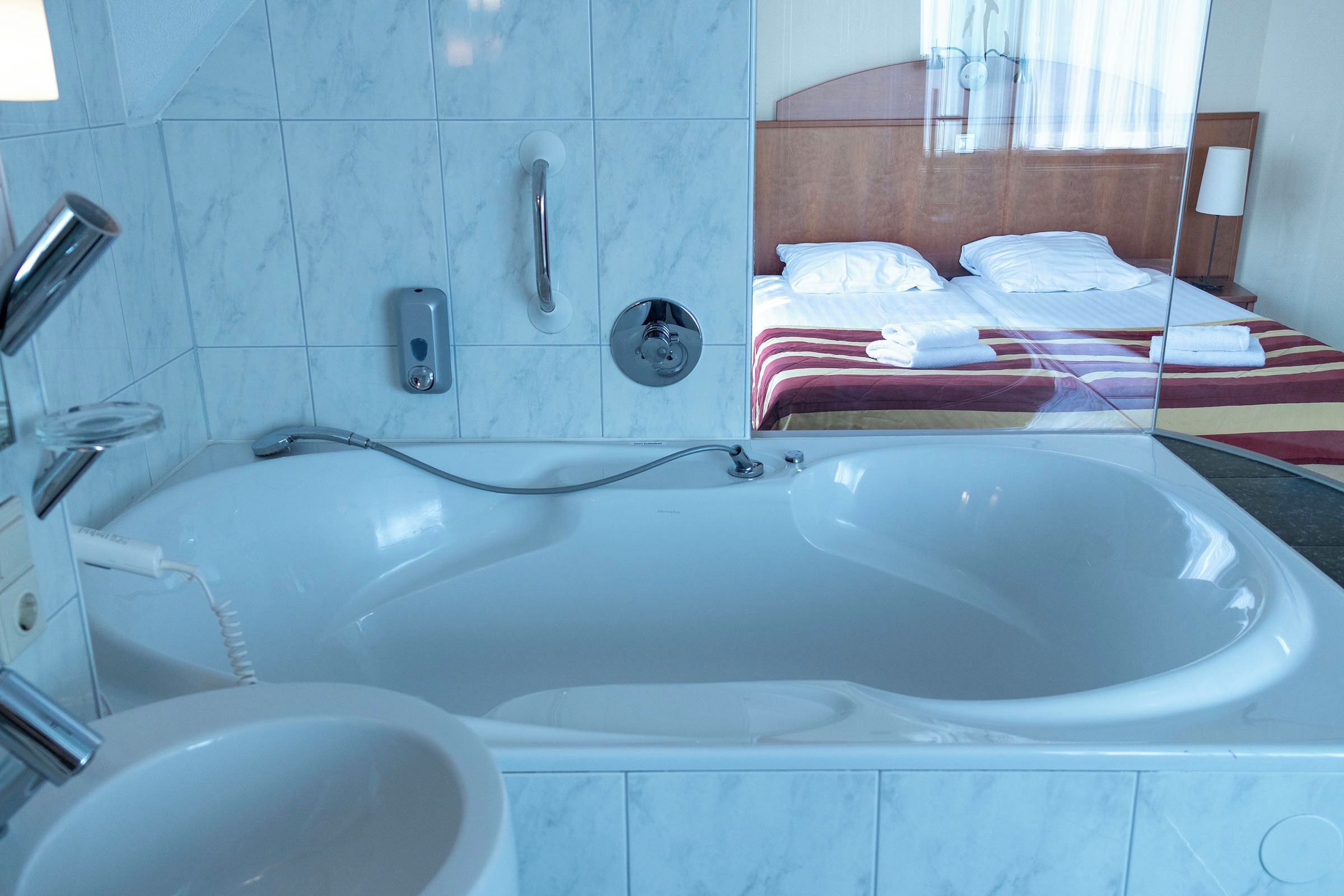 A modern bathroom with a large bathtub, a separate sink, and a mirror reflecting a neatly made bed with striped bedding.