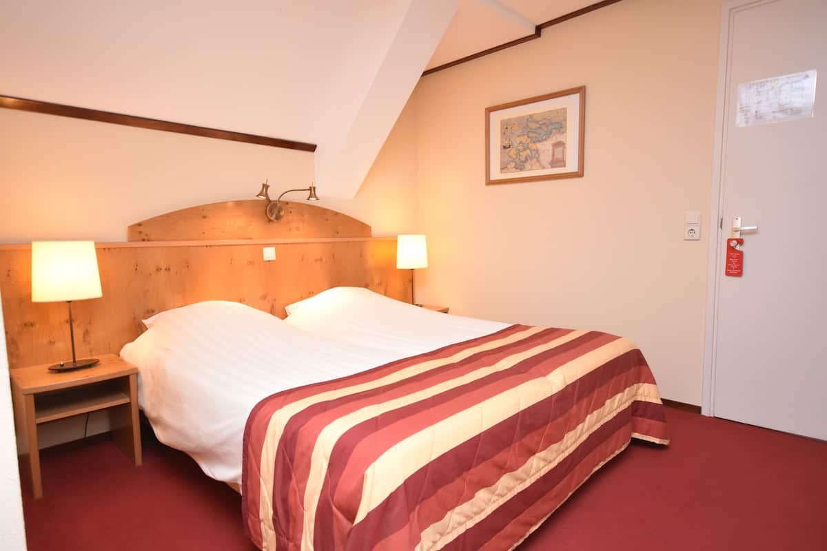 Une chambre d’hôtel avec un lit, deux lampes de chevet, une tête de lit en bois, un tableau encadré au mur et une porte avec un panneau rouge indiquant la sortie de secours.