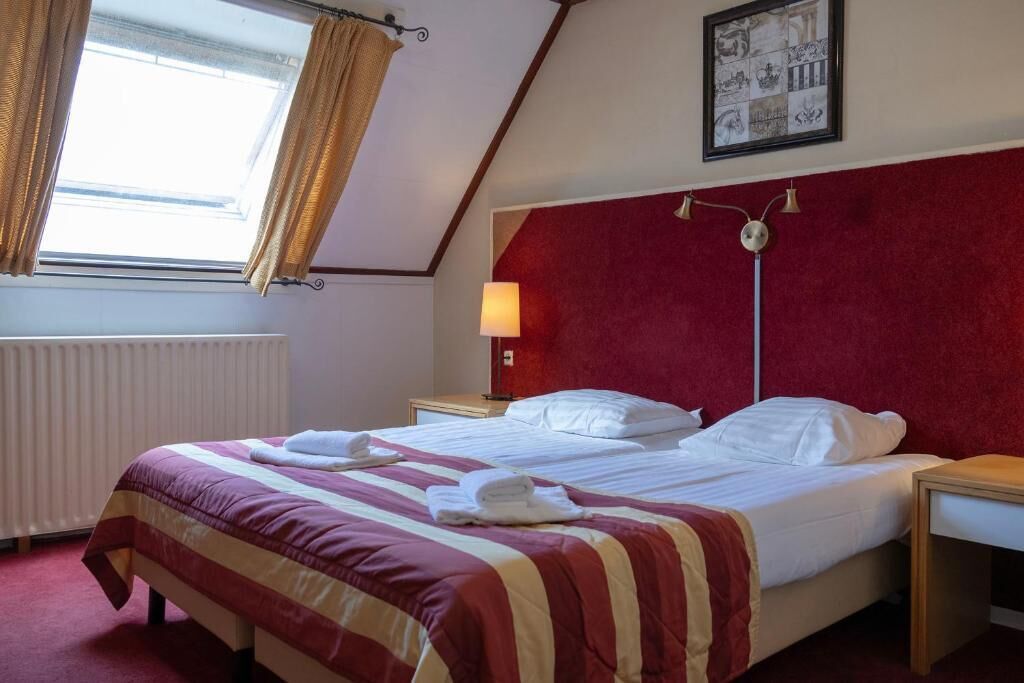 A hotel room with a bed, a nightstand, a lamp, a window with curtains, and a wall with a framed picture.