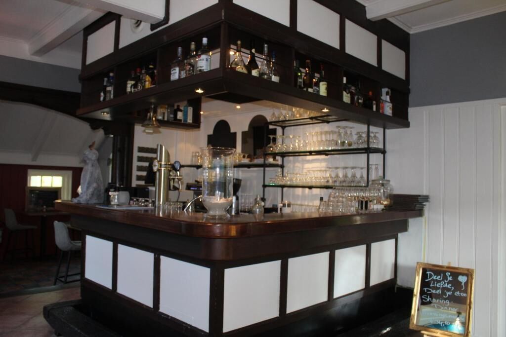 Una zona de bar con estantes llenos de botellas y vasos, una pizarra con escritura y una pequeña estatua.