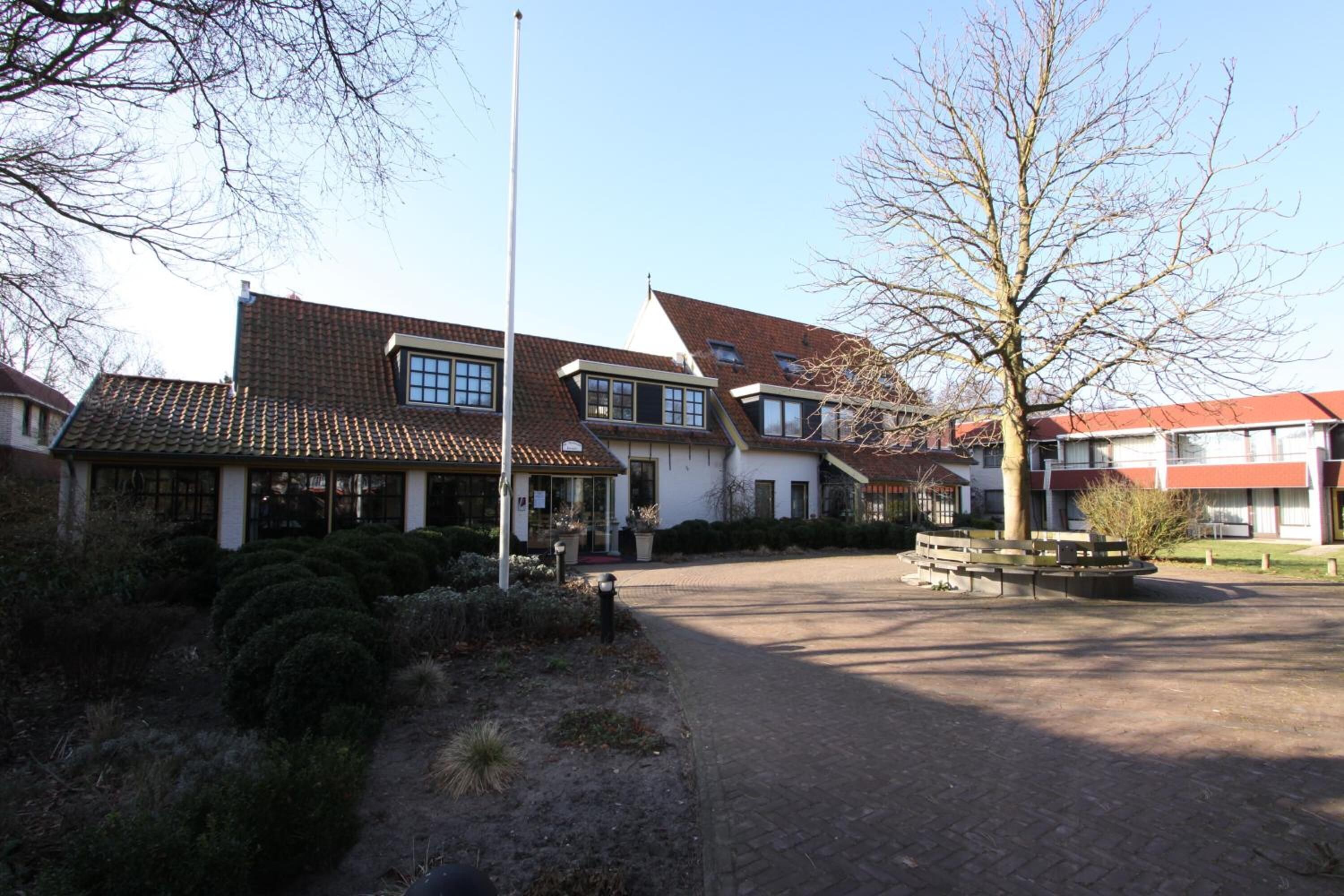 Un bâtiment doté d’un toit en tuiles, d’un mât pour drapeau et d’une cour pavée avec des bancs.