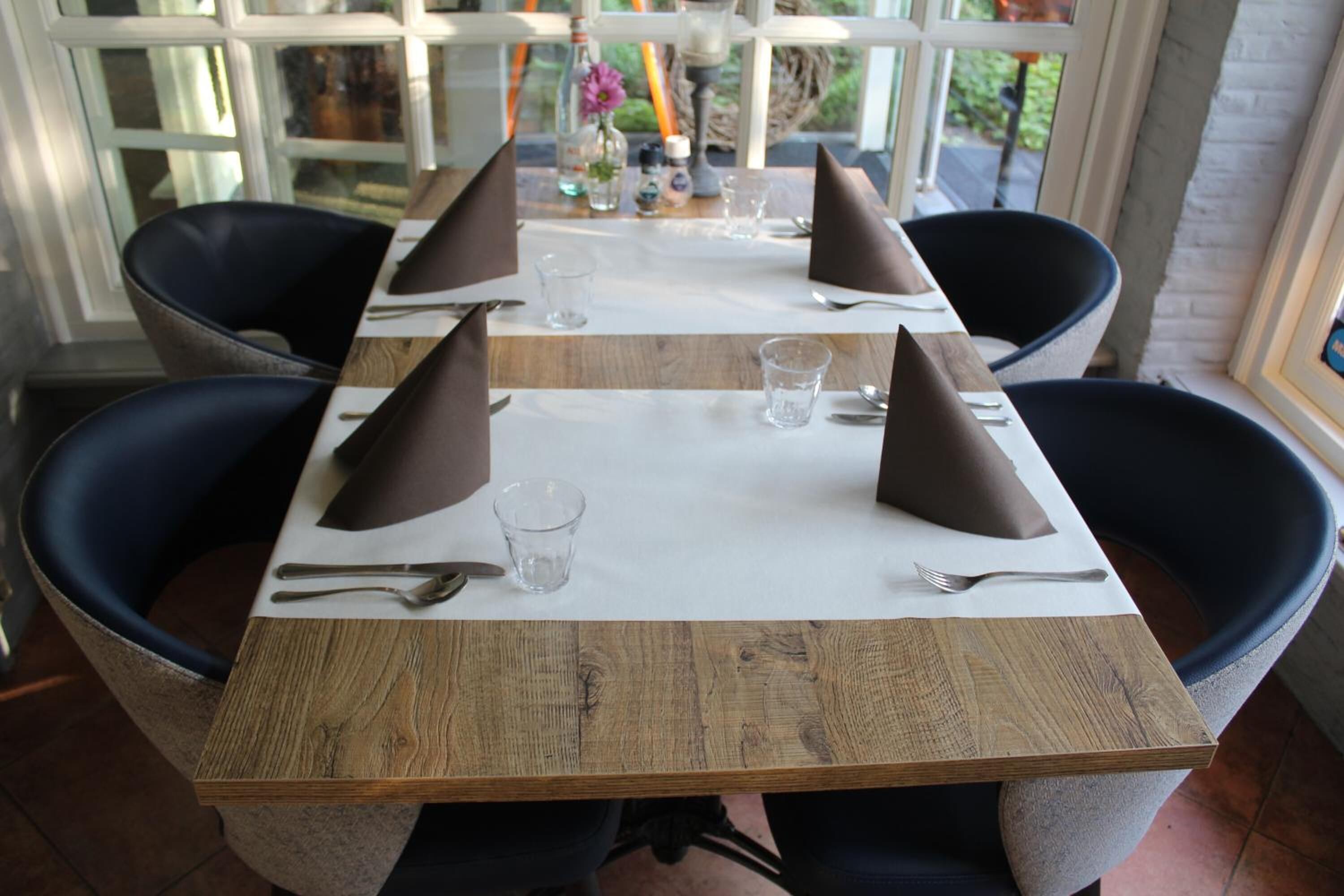Ein Esstisch ist mit vier Stühlen gedeckt, jedes mit einer Serviette, Besteck und einem Glas. Der Tisch ist aus Holz und mit einer weißen Tischdecke bedeckt.