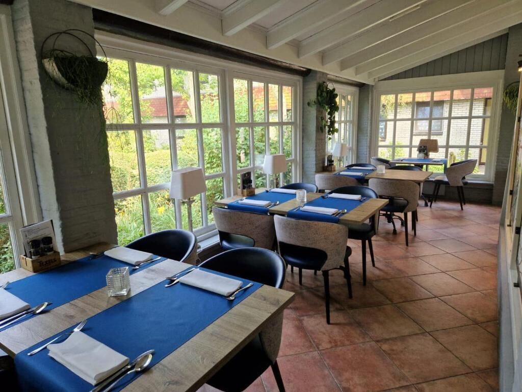 A dining area with tables set for four, large windows, and a view of greenery outside.