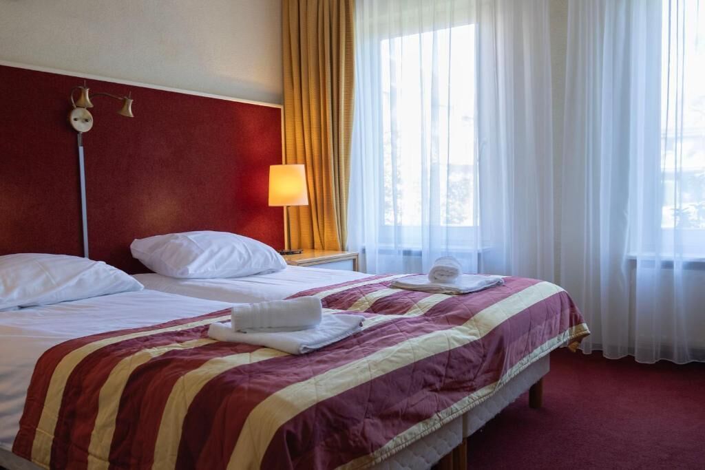 Quarto de hotel com cama, mesa de cabeceira, abajur e janela com cortinas.