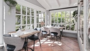 Une salle à manger lumineuse, avec de grandes fenêtres, des murs blancs et un sol carrelé.