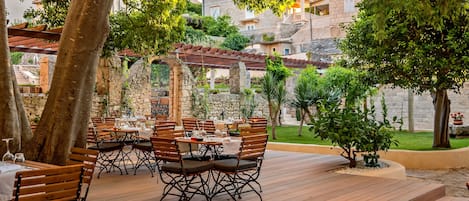 An outdoor dining area with wooden tables and chairs, surrounded by trees and stone walls.