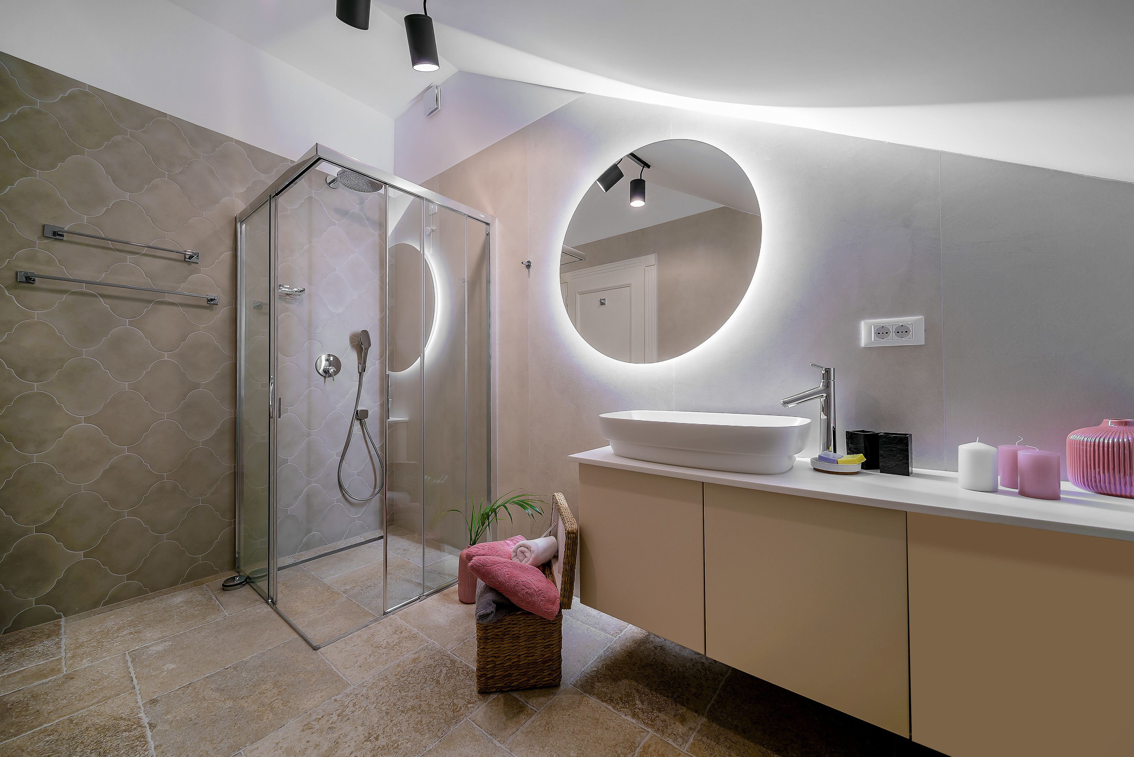 A modern bathroom with a glass shower enclosure, a round mirror, a white sink, and a vanity with a basket of towels.