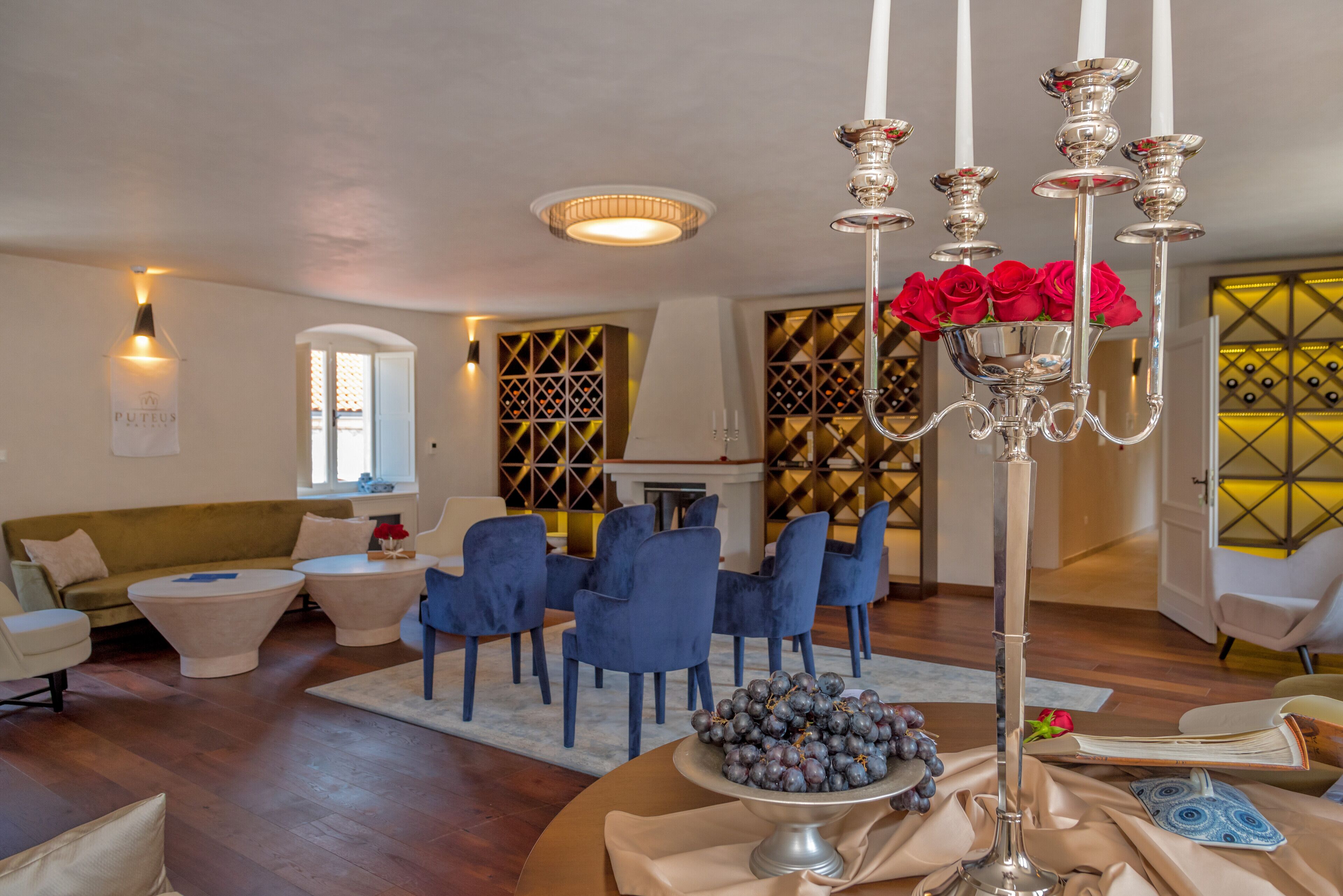 Een moderne woonkamer met een kroonluchter, blauwe stoelen, een ronde tafel met een schaal druiven en een bank.