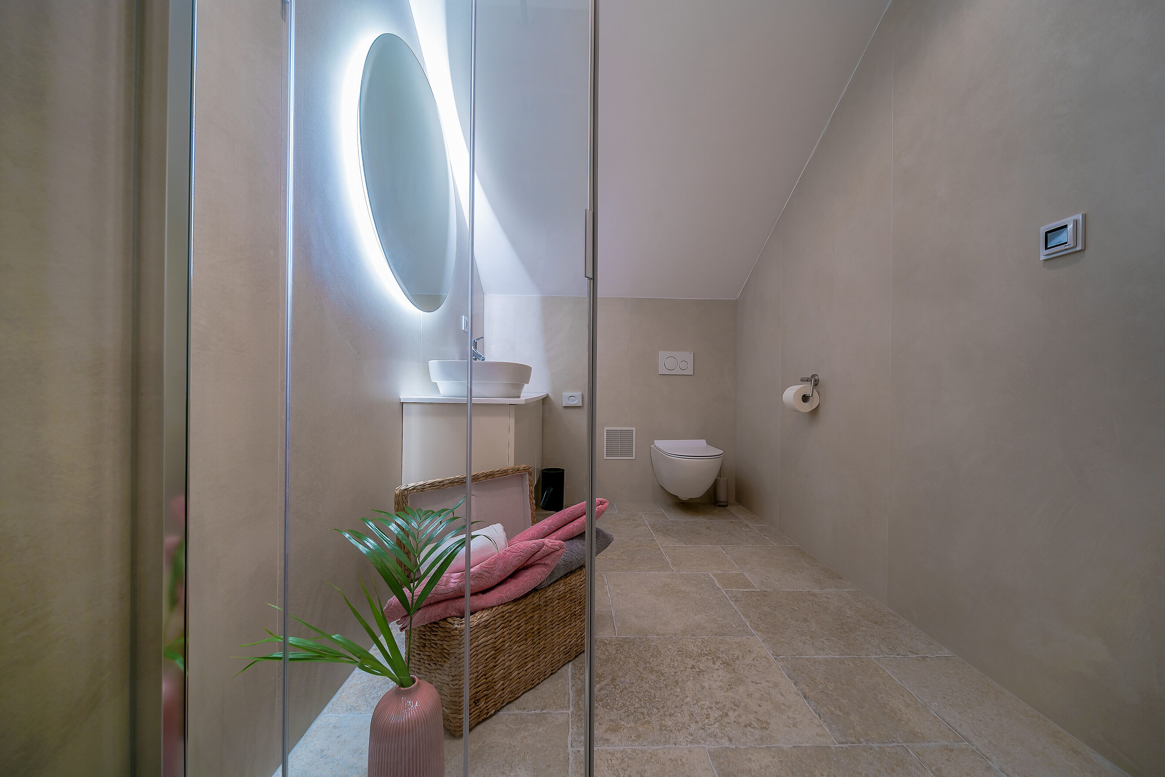 Une salle de bain moderne avec une cloison en verre, un grand miroir, un lavabo, des toilettes et un panier en osier rempli de serviettes.