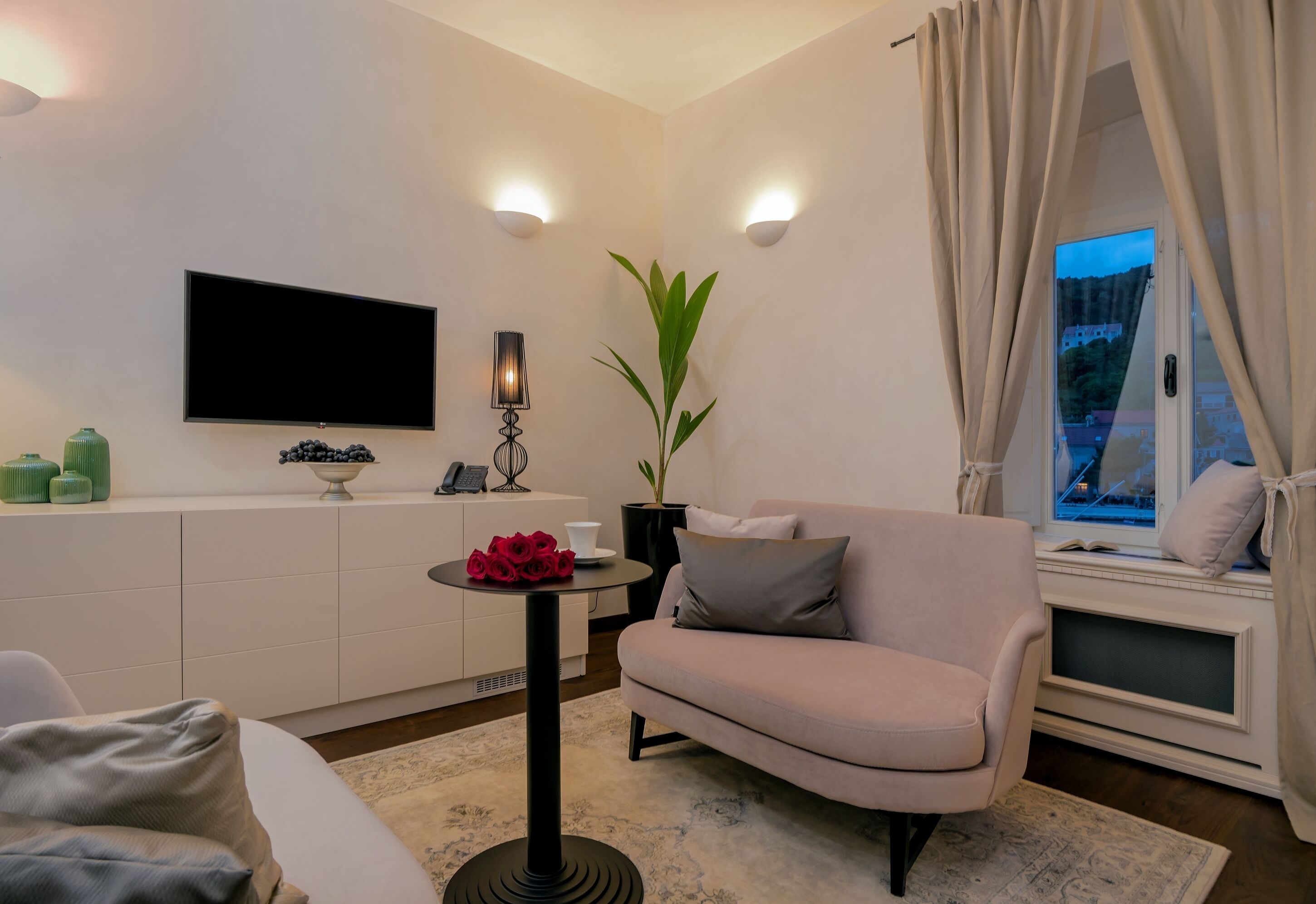 A modern living room with a sofa, a small table with a vase of red roses, a potted plant, and a flat-screen TV mounted on the wall.