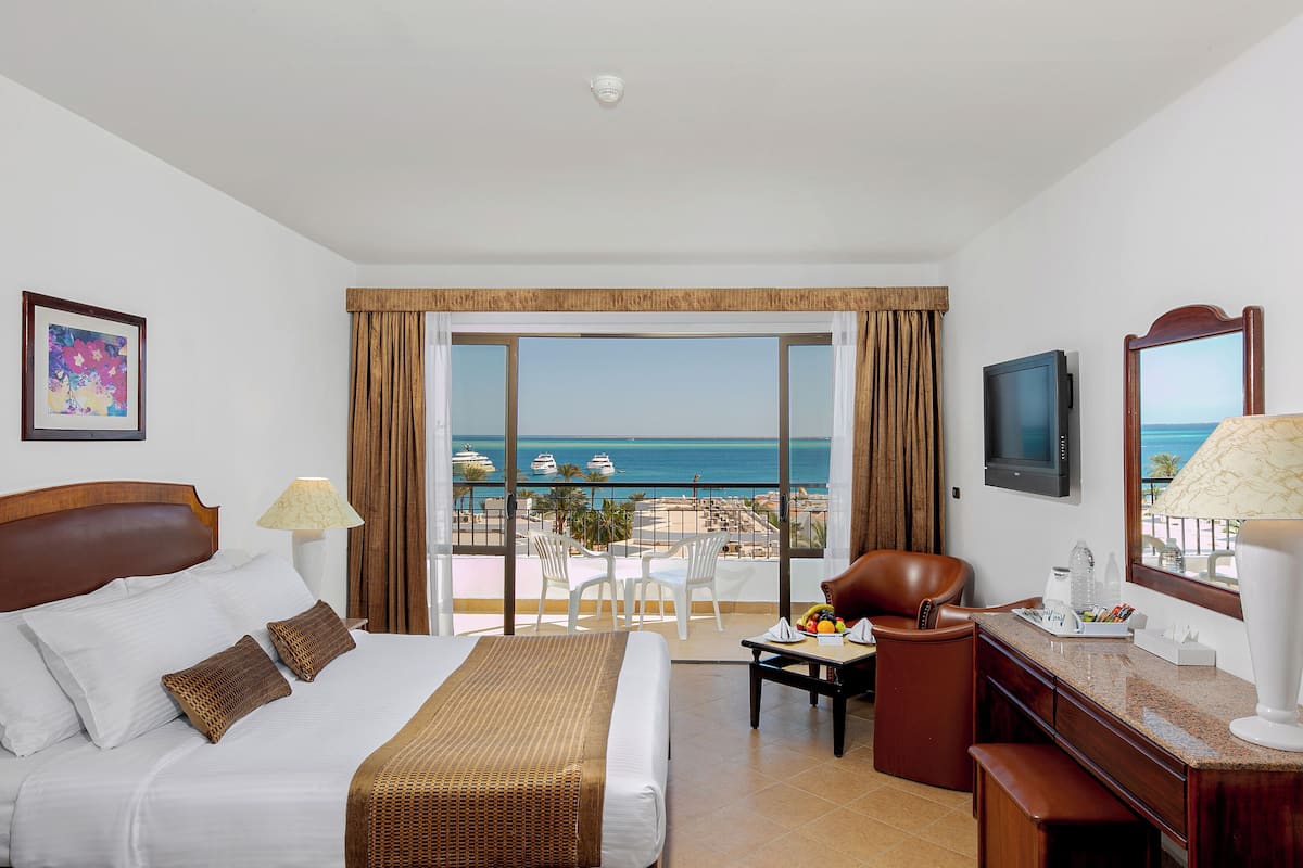 A hotel room with a bed, a desk, a chair, a TV, and a balcony with a view of the ocean.
