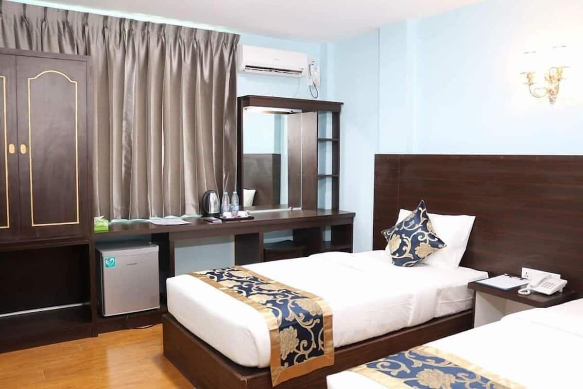 A hotel room with a bed, bedside table, and a desk with a chair. There is a small refrigerator, a mirror, and a window with curtains.