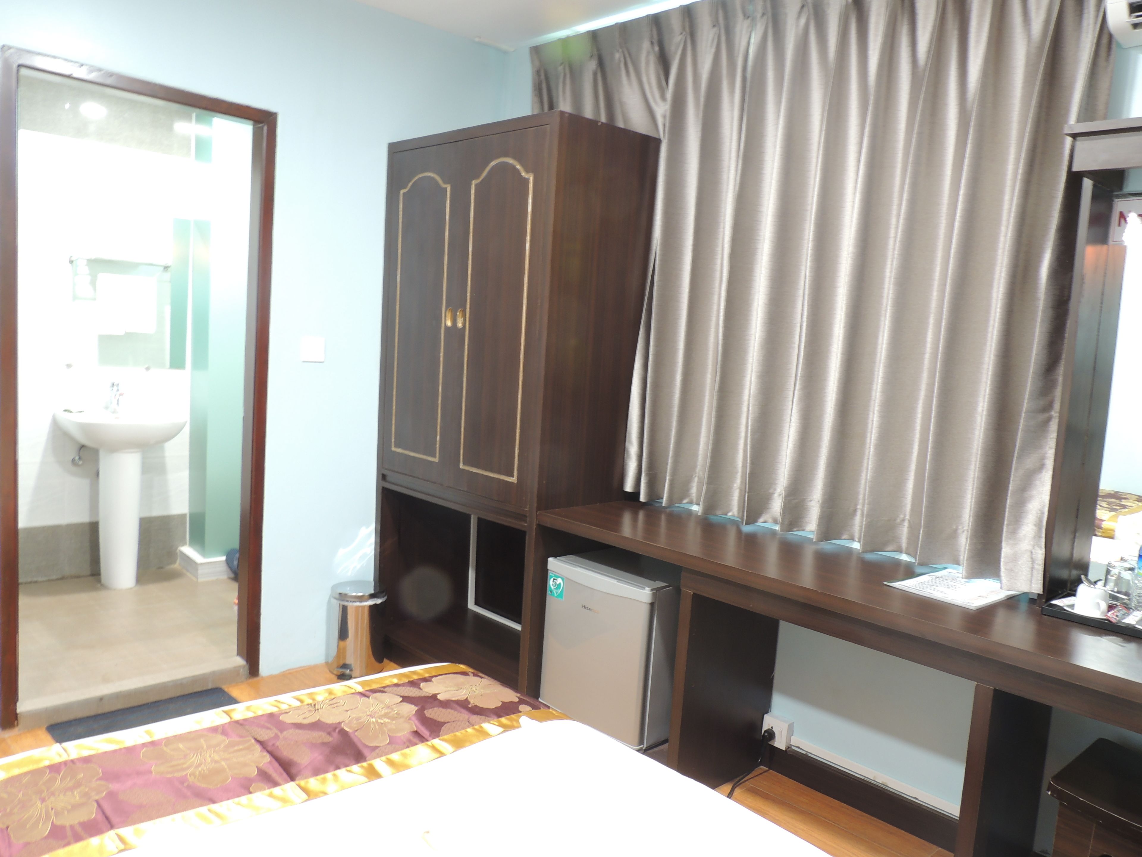 Una habitación de hotel con cama, armario, refrigerador pequeño, lavabo y ventana con cortinas.