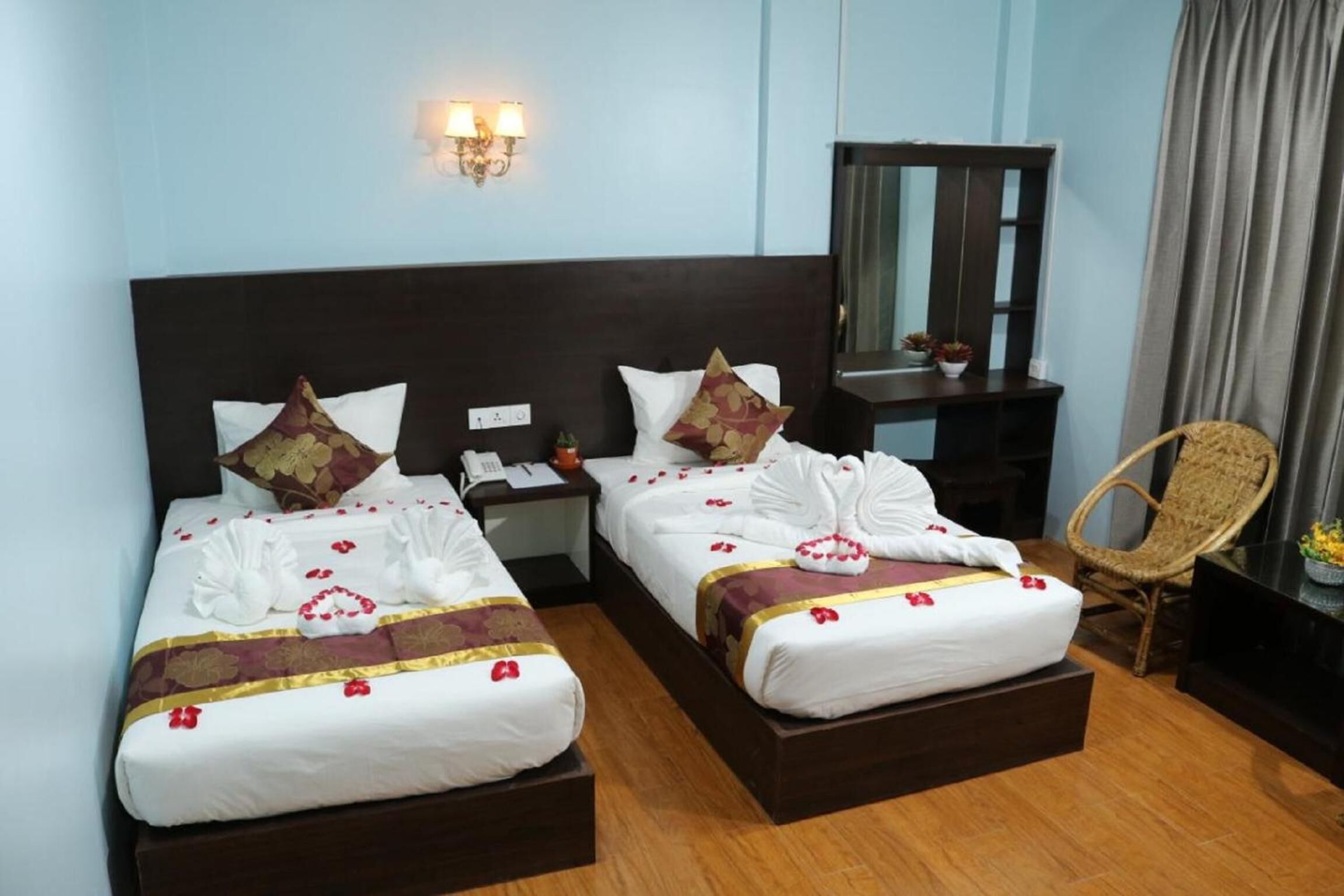 A hotel room with two beds, each with white linens and red floral accents, a wooden headboard, bedside tables with lamps, a wicker chair, and a mirror on a dresser.