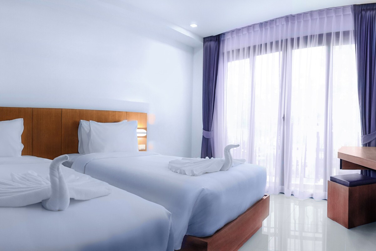 Ein Hotelzimmer mit zwei Betten, einem hölzernen Kopfteil, einem Schreibtisch und einem großen Fenster mit Vorhängen.