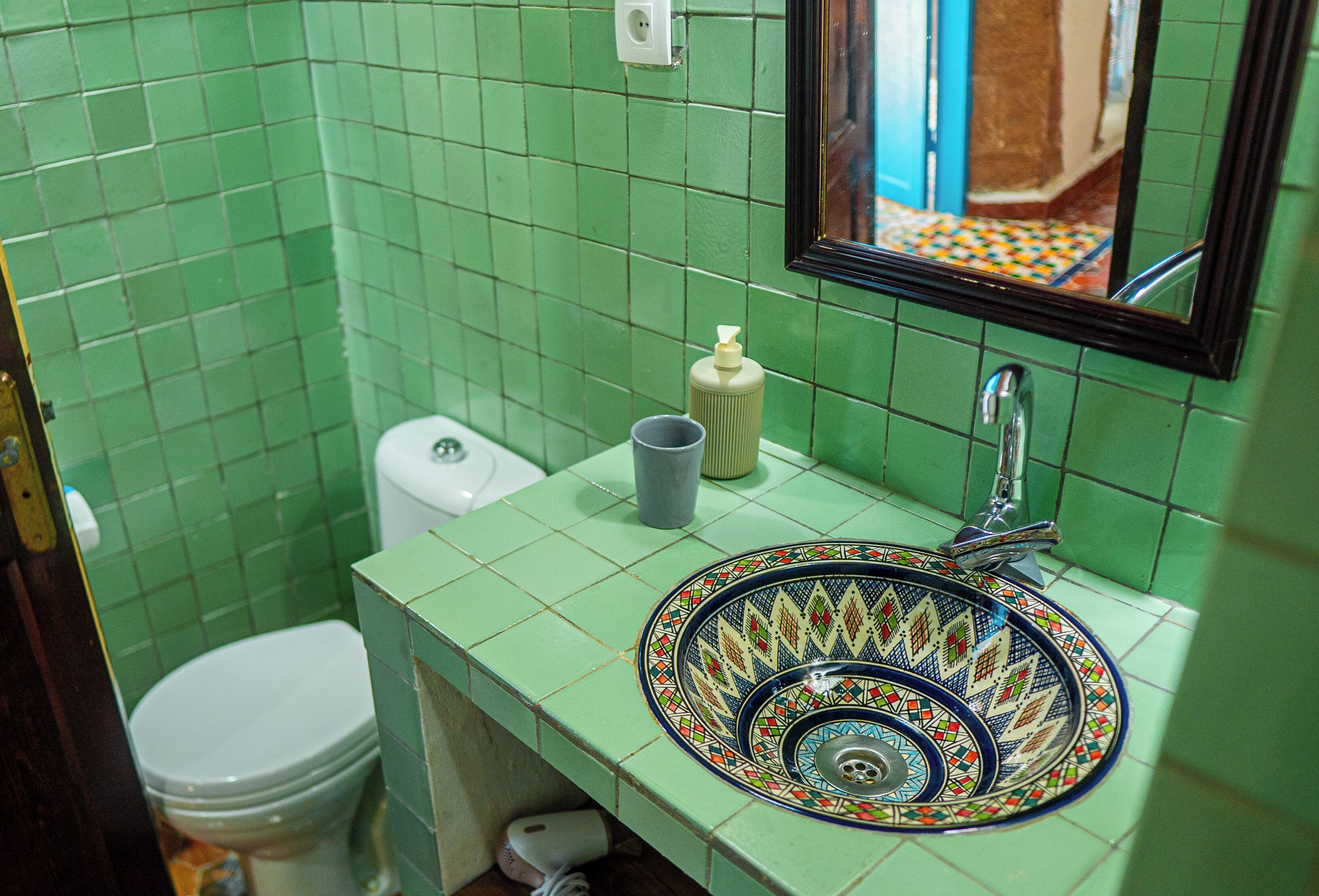 Ein Badezimmer mit grünen Fliesen, einer Toilette, einem Waschbecken mit dekorativer Schale und einem Spiegel.