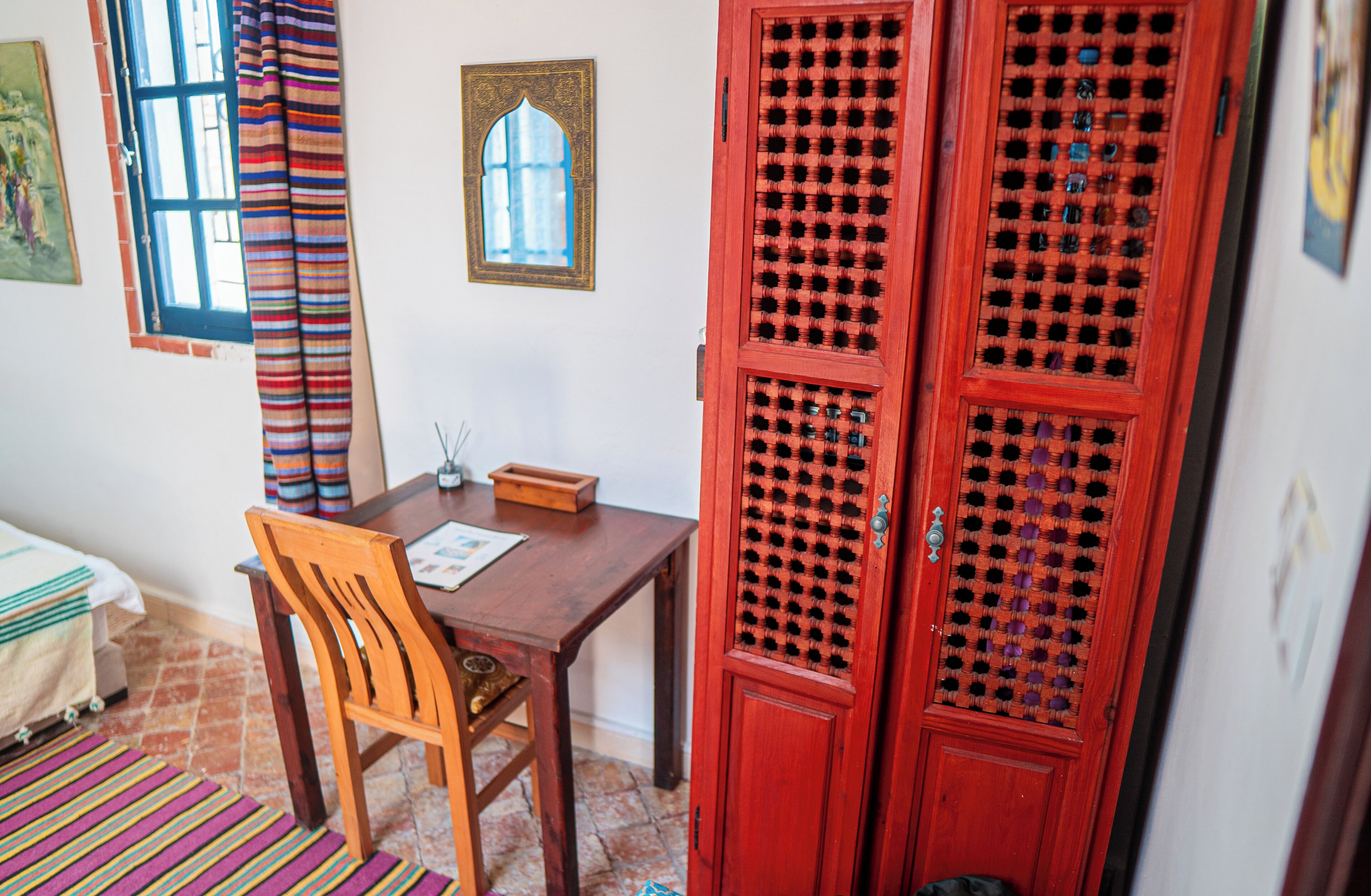 A room with a wooden table, a chair, a red door with a lattice design, a window with curtains, and a small wooden box on the table.