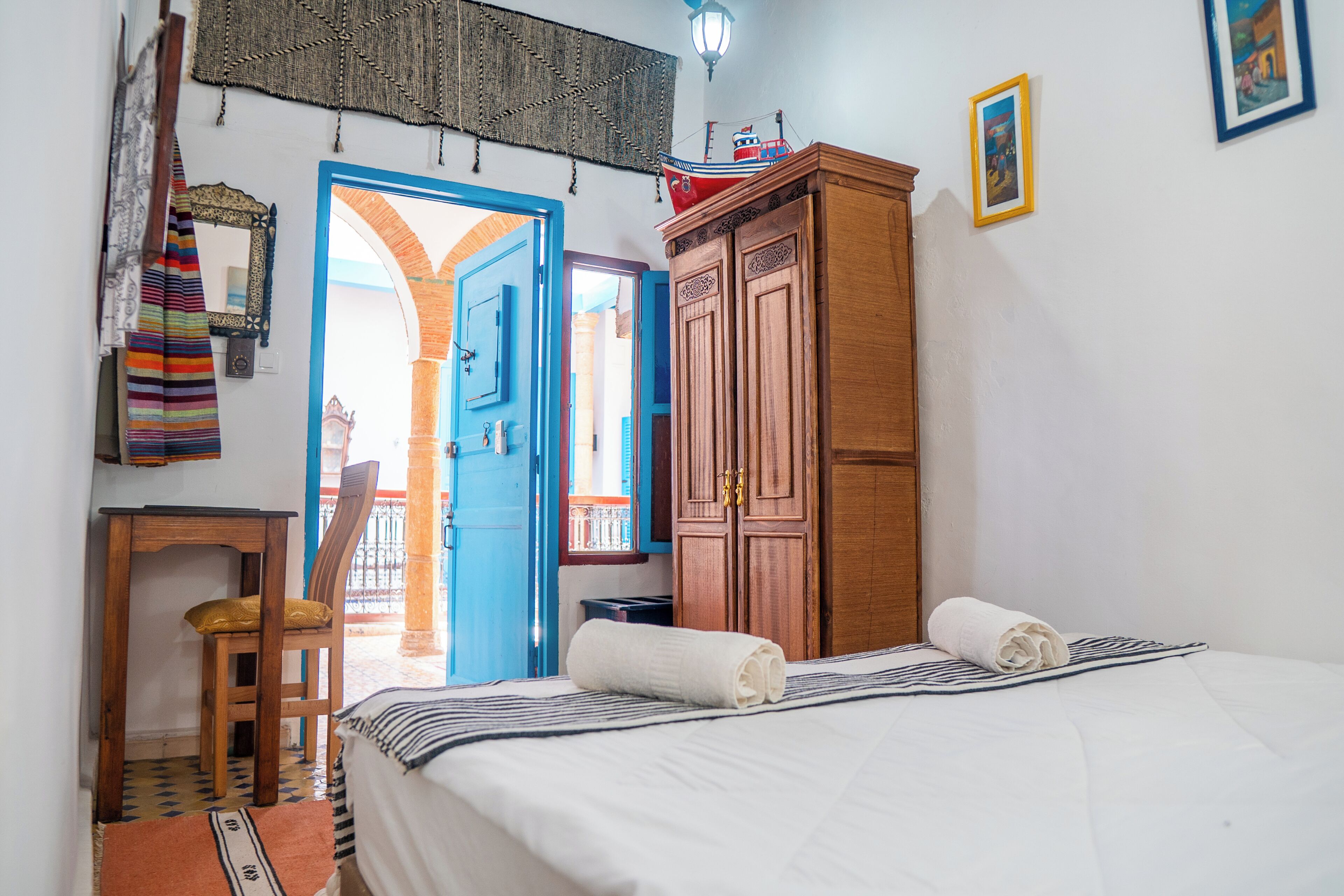 A bedroom with a bed, a wooden wardrobe, a chair, a small table, and a doorway leading to an outdoor area.