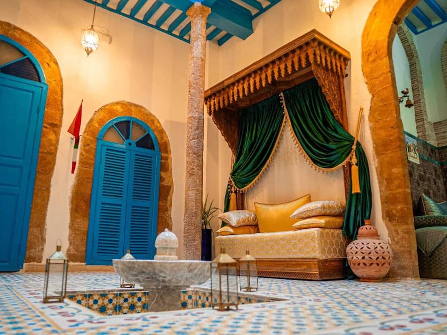 Una habitación con una puerta azul, una cama de madera con cortinas verdes, piso de baldosas y un pilar de piedra.