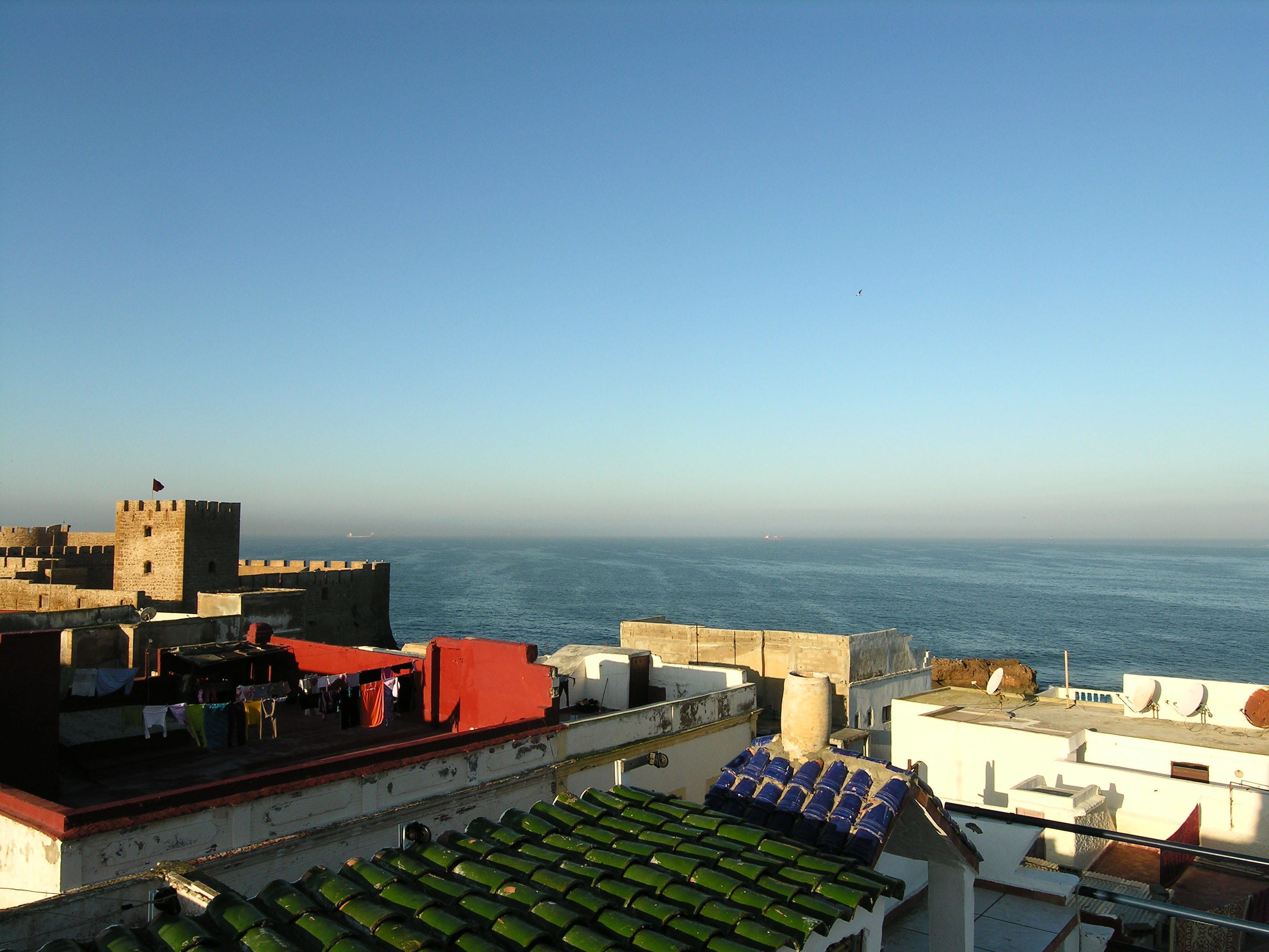 A coastal cityscape with a prominent tower, terracotta rooftops, and a clear view of the sea.