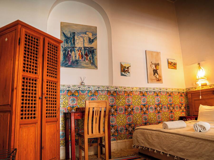 Een kamer met een bed, een houten garderobe, een stoel, een betegelde muur en kunst aan de muur.